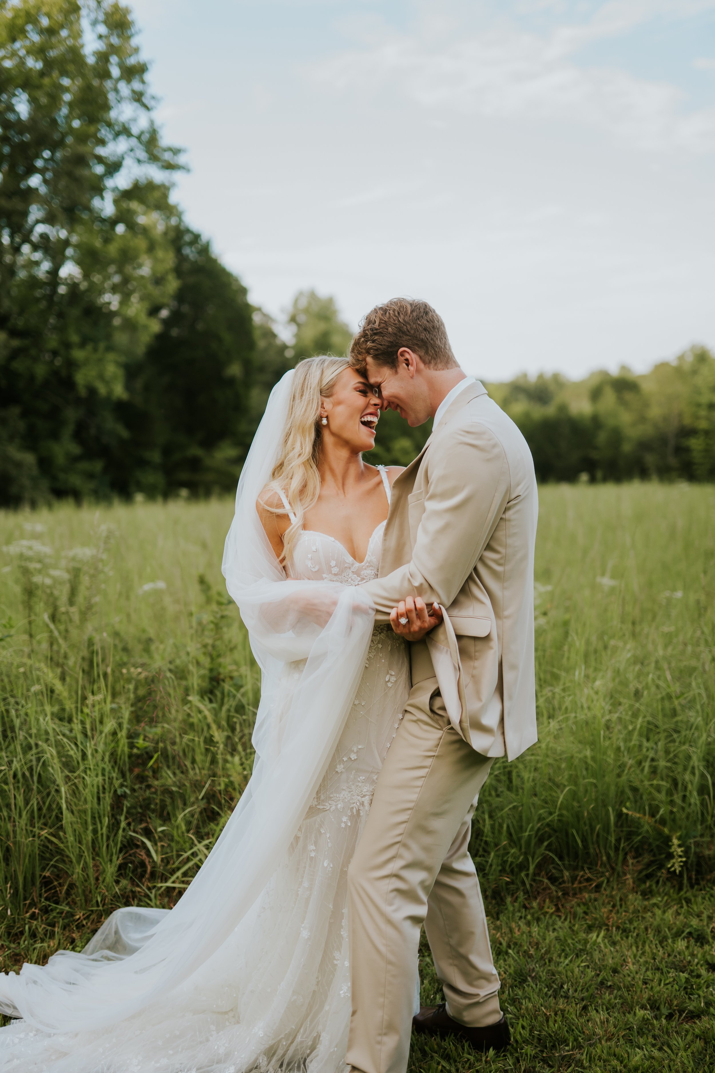 A bride and groom share a joyful moment outdoors in a green field with trees in the background.