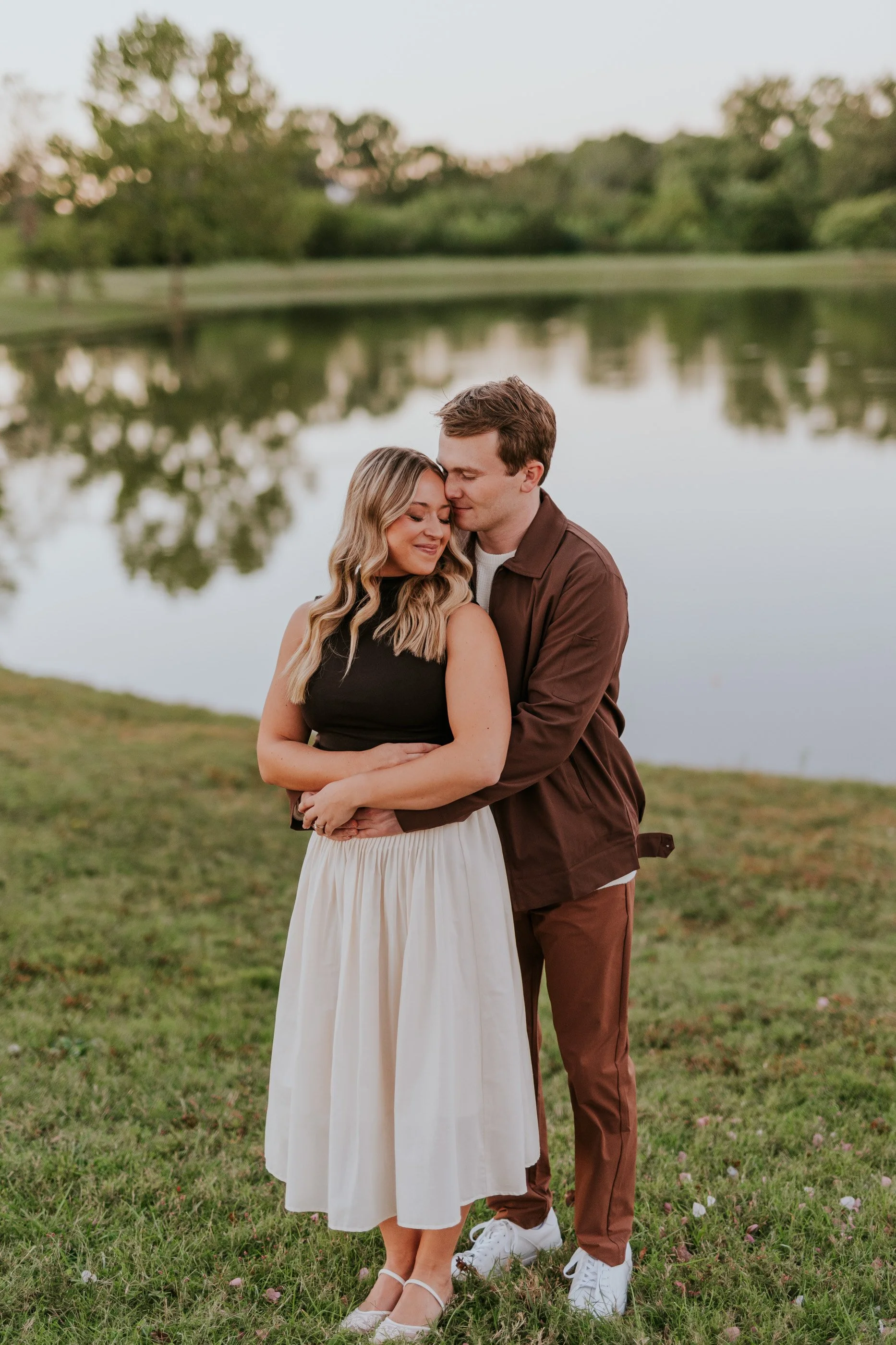A young couple embracing near a lake at sunset, with trees reflected in the water behind them.