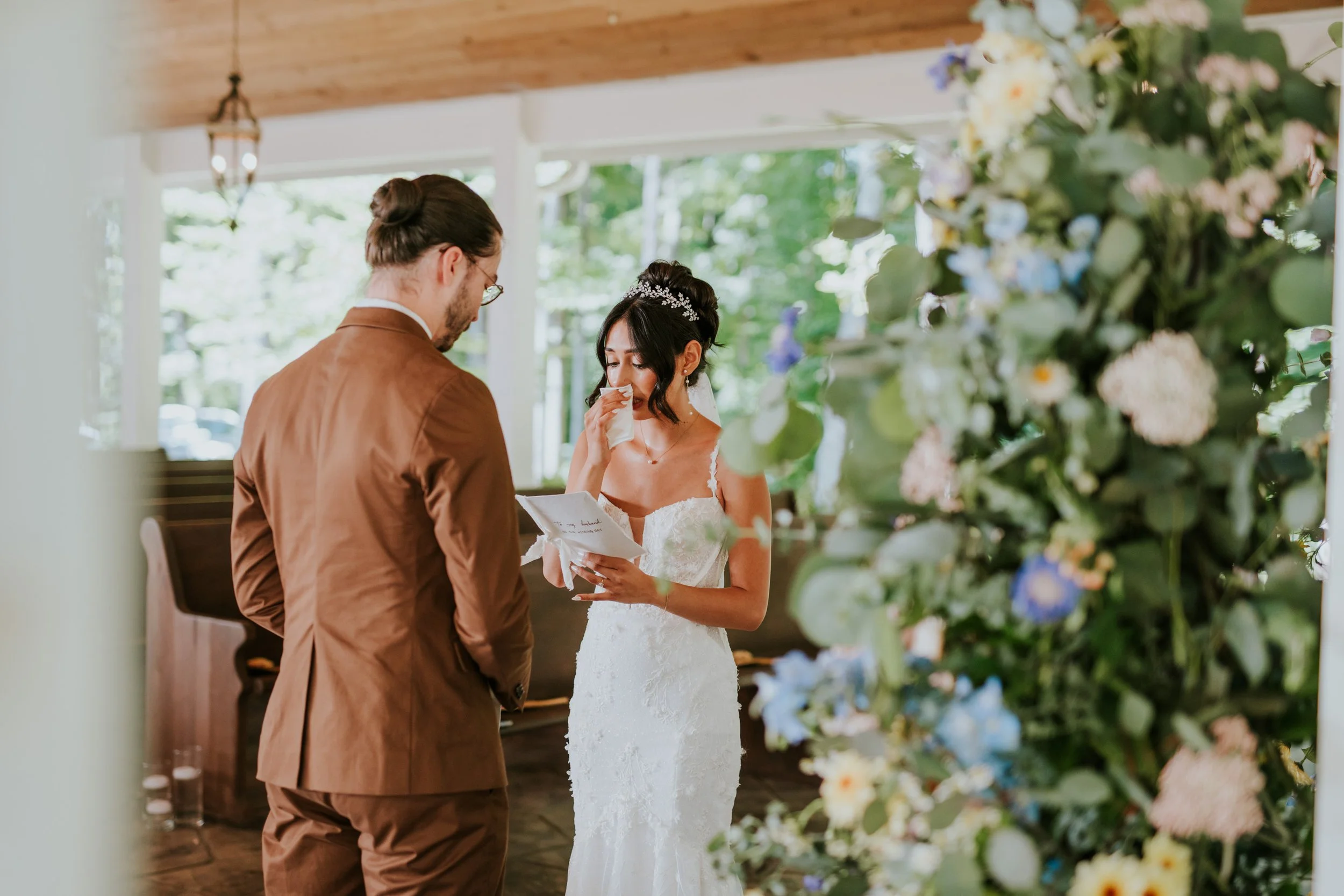 A bride and groom during their wedding ceremony, with the bride emotional and holding a tissue, and the groom standing with his back to the camera, in a brightly lit indoor setting with large windows and floral decorations.