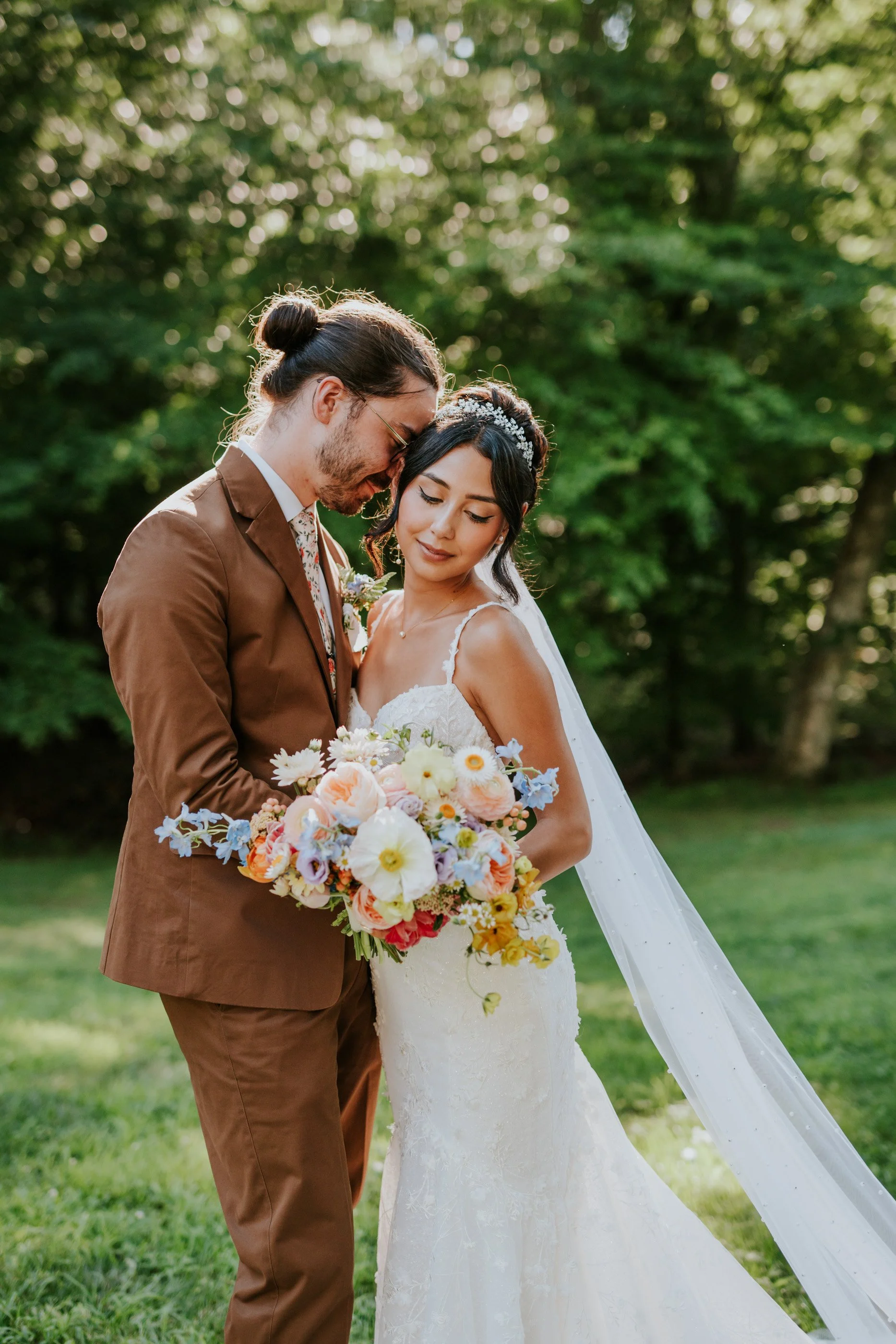 A bride and groom stand close together outdoors, the groom leaning his forehead against the bride's temple. The bride is holding a colorful bouquet of flowers and is dressed in a white lace wedding gown. The groom wears a brown suit with a floral tie