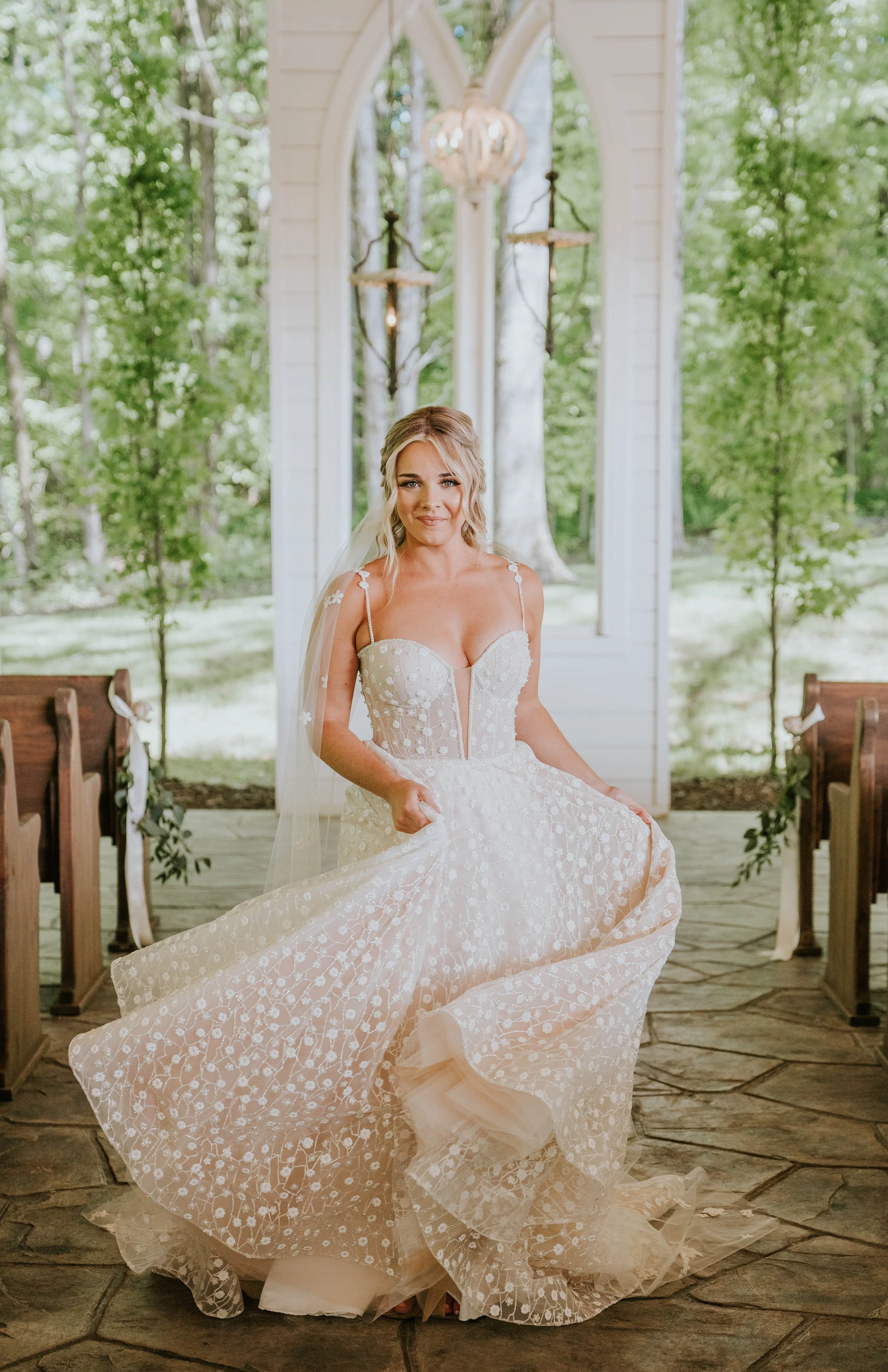 A bride in a wedding dress holding the hem inside a church with a wooden floor, white walls, sunlight, and green trees outside.