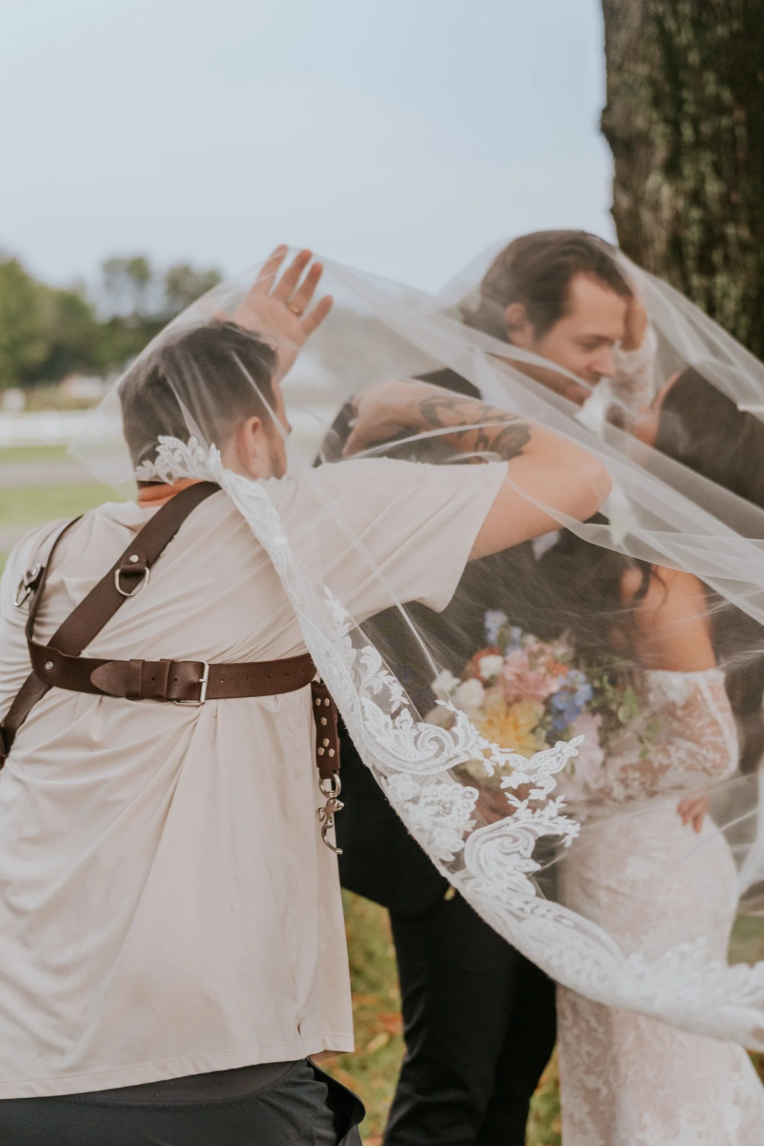 Two people, one in a wedding dress, are covered by a veil held by a person in a beige shirt and suspenders. The scene appears to be outdoors near a tree.