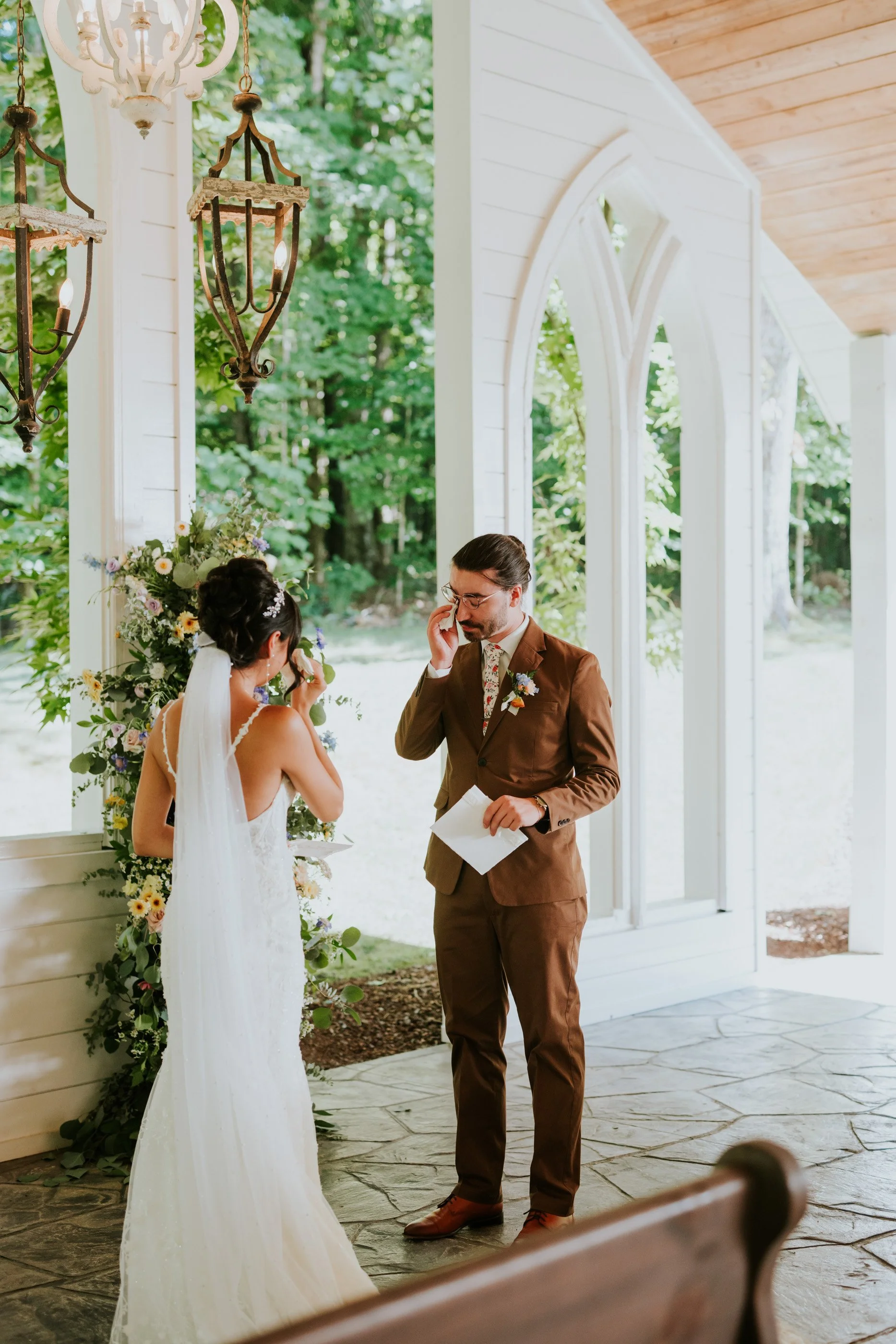 A bride and groom during their outdoor wedding ceremony inside a white chapel with large arched windows. The bride is wiping tears, and the groom, holding a piece of paper, is talking to her. There are hanging lanterns and a floral arrangement in the