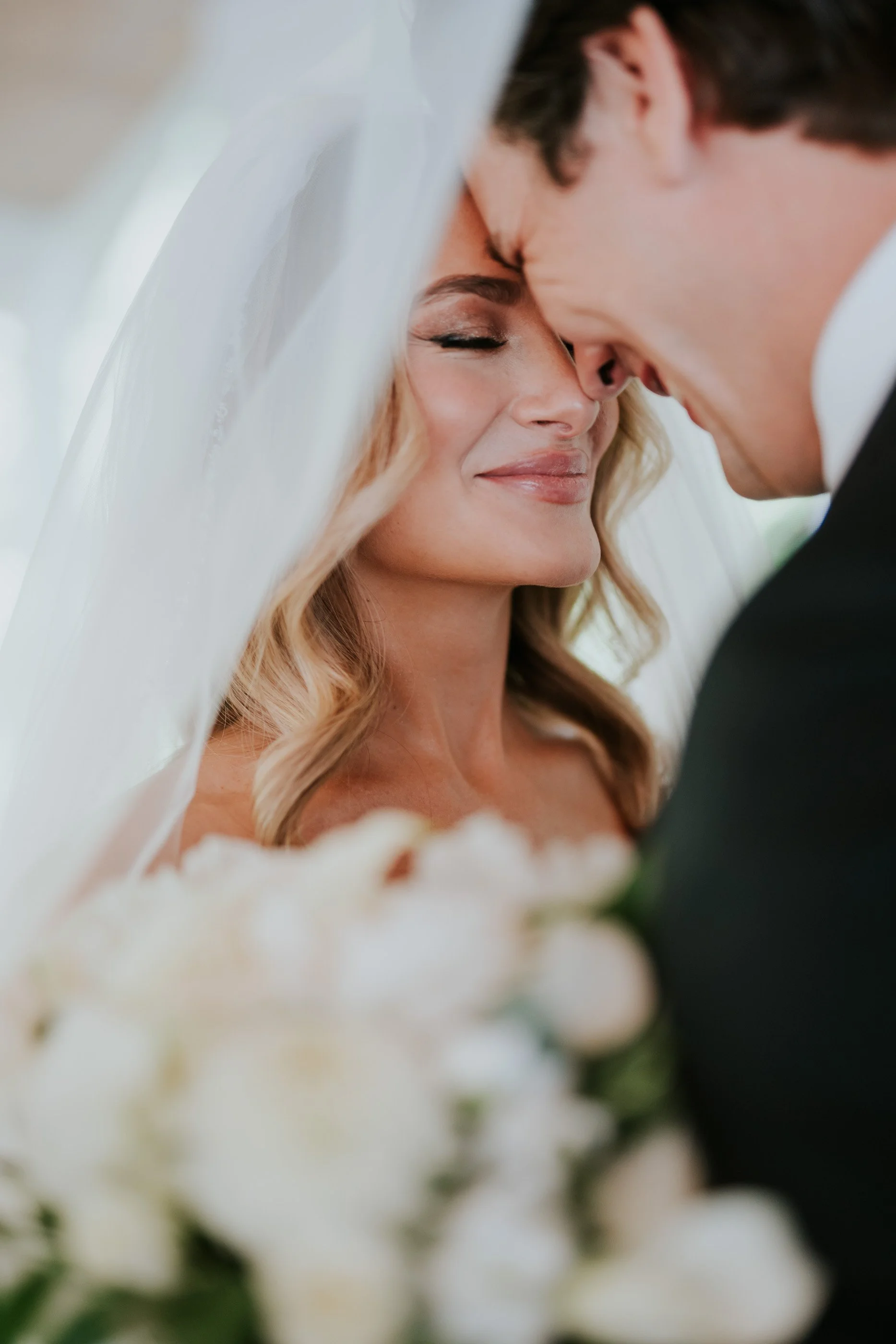 Close-up of a bride and groom with foreheads touching, smiling gently during their wedding ceremony, with a white veil and bouquet visible.