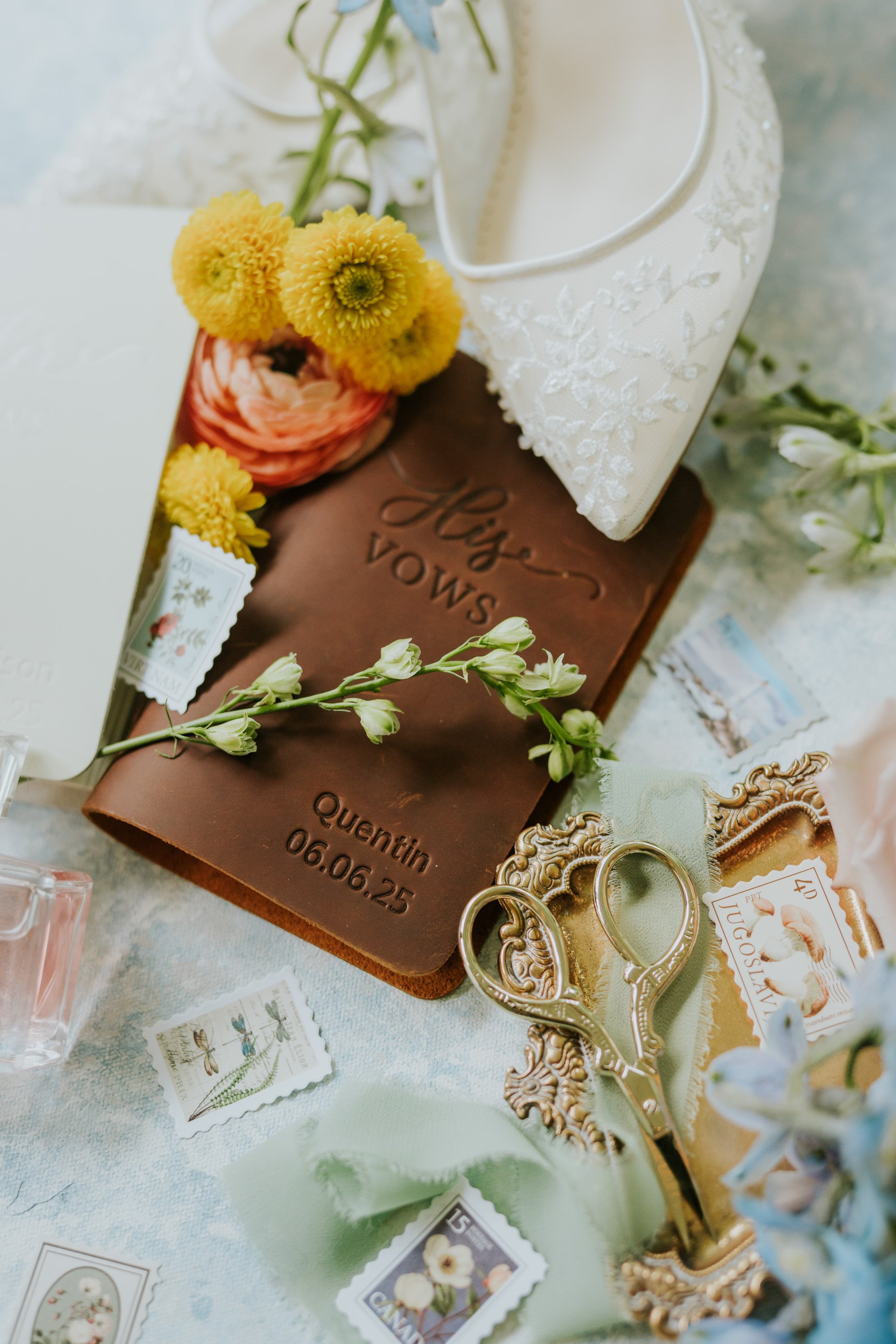 A wedding invitation suite with a chocolate leather cover engraved with 'Les Vows' and the name 'Quentin' with the date '06.06.25'. The setup includes a small bouquet of yellow flowers, a pair of ornate gold wedding rings, vintage-style postage stamp