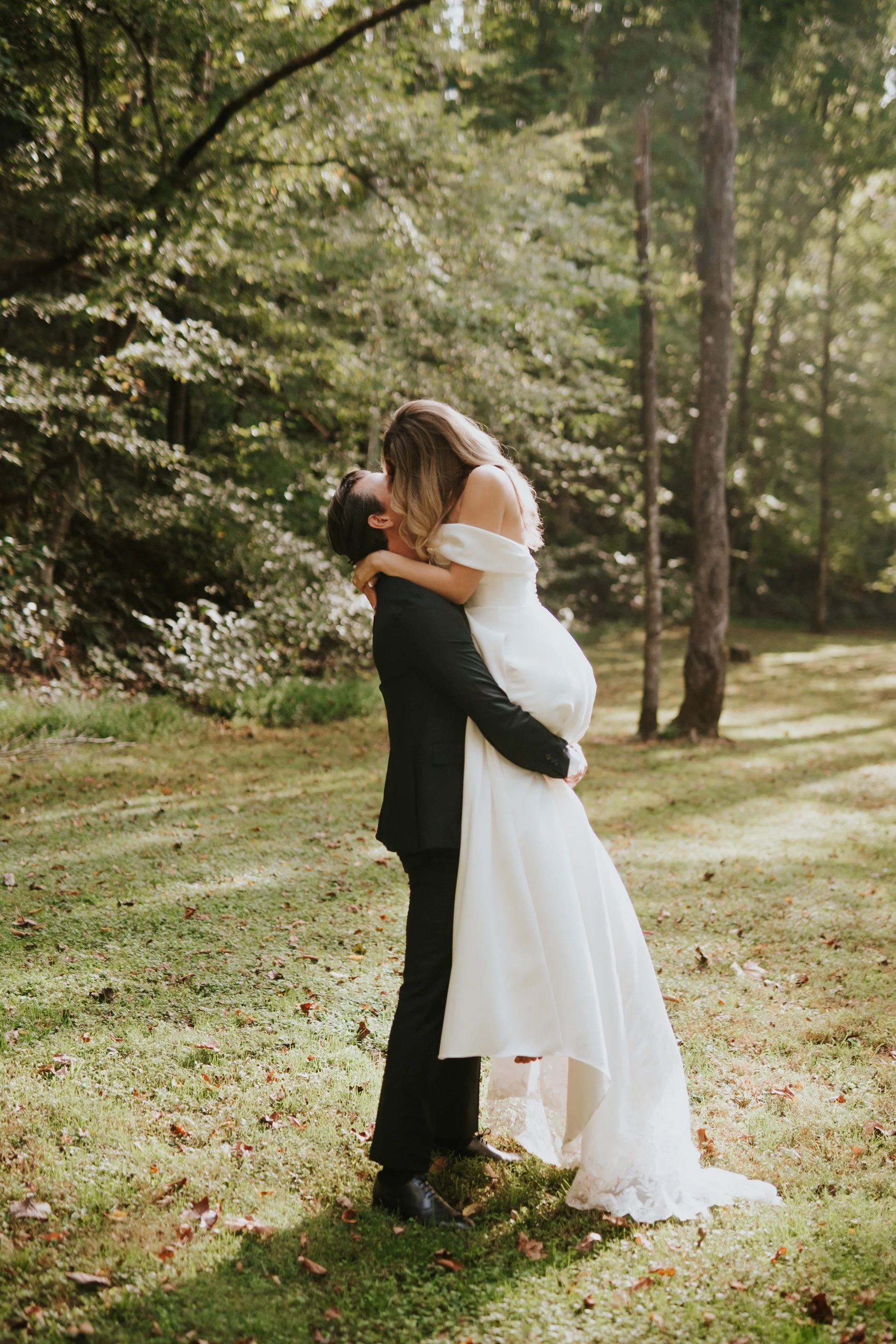 A couple on their wedding day, with the groom lifting the bride in a forested outdoor setting, sunlight filtering through the trees.