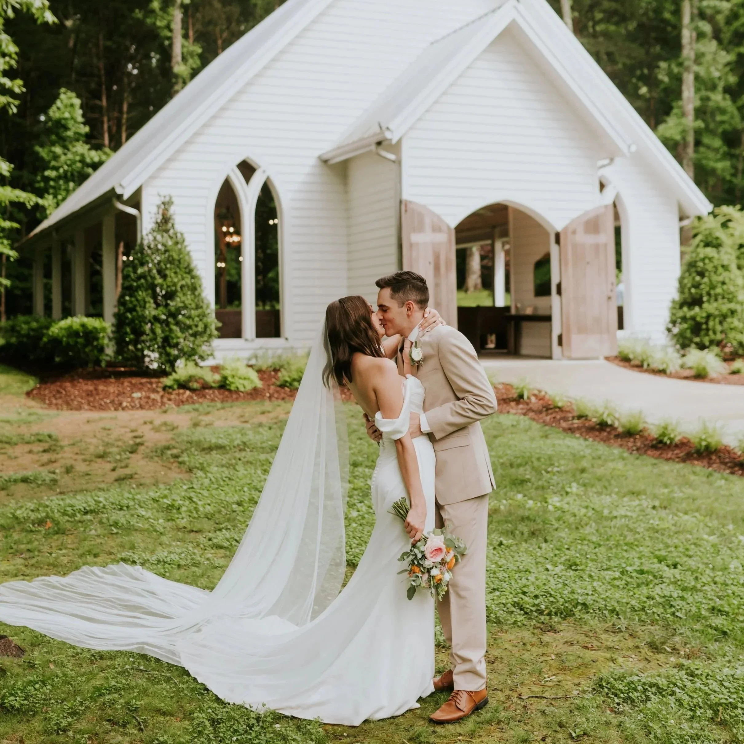 A bride and groom kiss in front of a white chapel with open wooden doors, surrounded by greenery on a lawn.