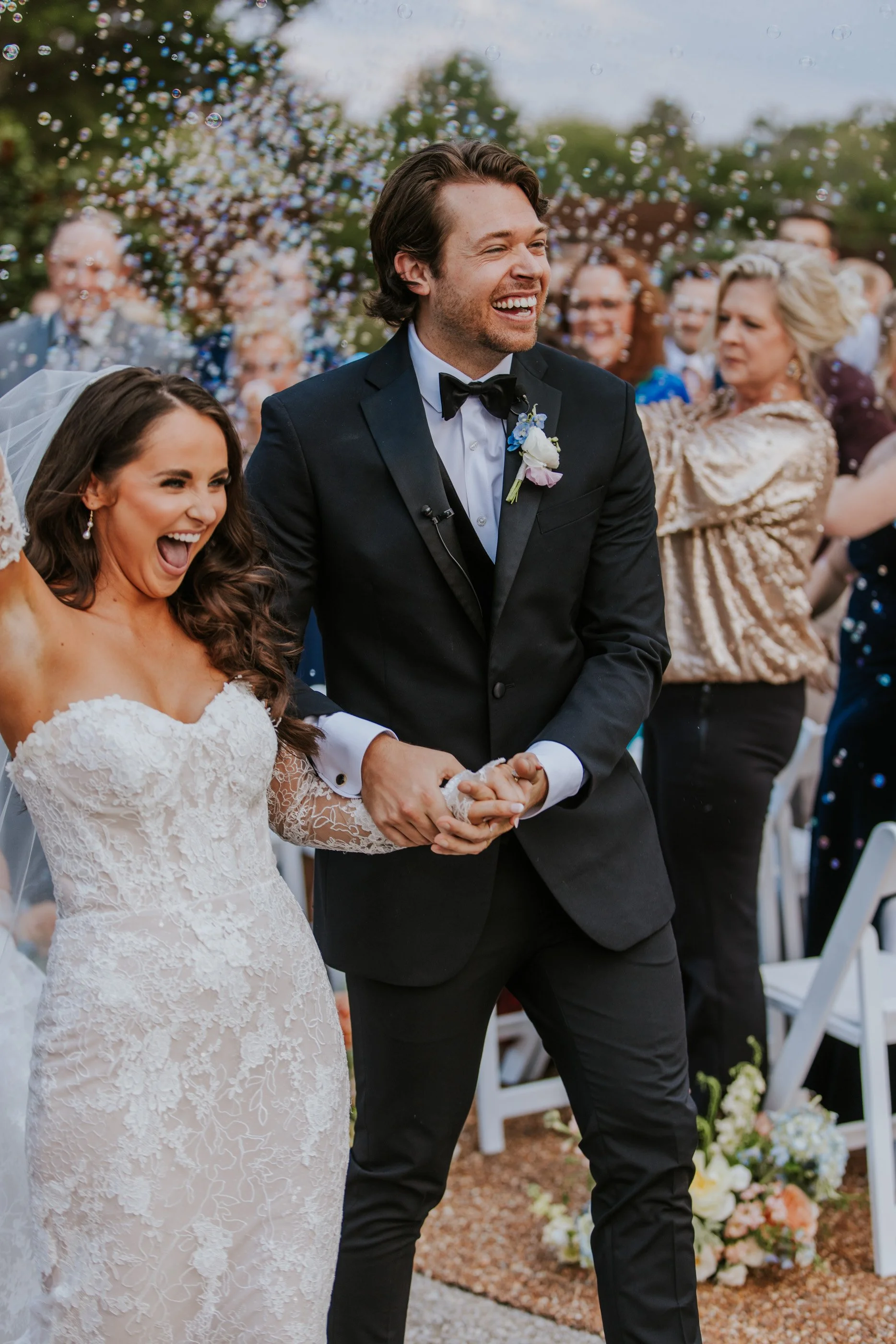 Happy wedding couple holding hands during the ceremony, surrounded by cheerful guests, outdoors.