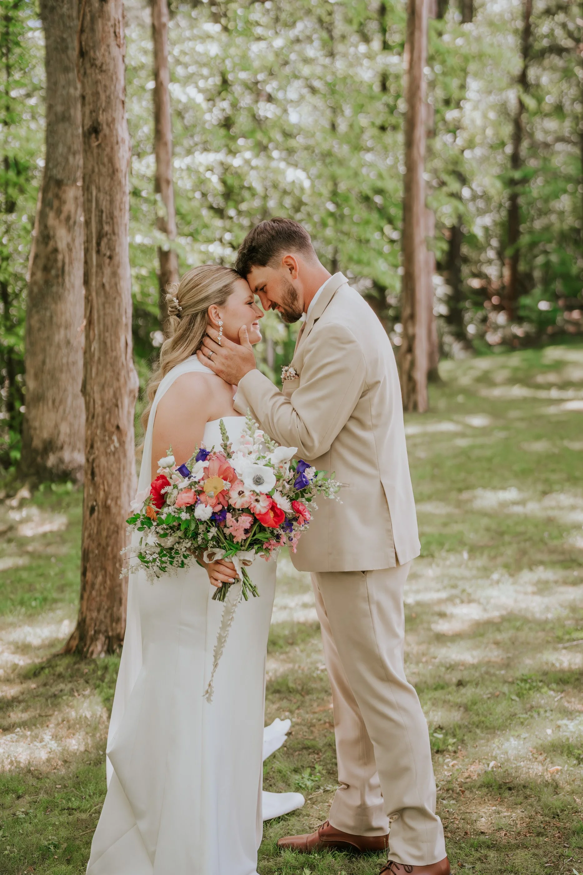 A bride and groom sharing a tender moment outdoors among trees during their wedding, with the groom gently holding the bride's face and both smiling softly.