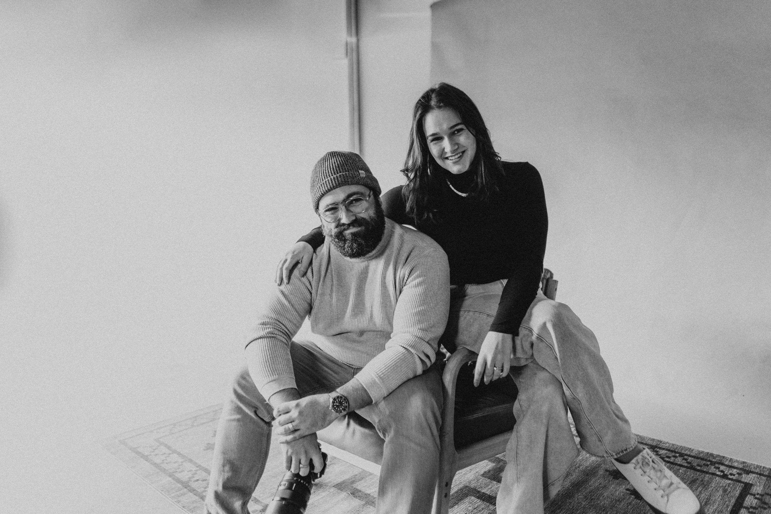A black-and-white photo of a man and a woman seated on a wooden chair. The man has a beard, glasses, wearing a beanie, sweater, and jeans, with a camera in hand. The woman has shoulder-length hair, wearing a black top, light-colored pants, and white sneakers, smiling with her arm around the man's shoulders.