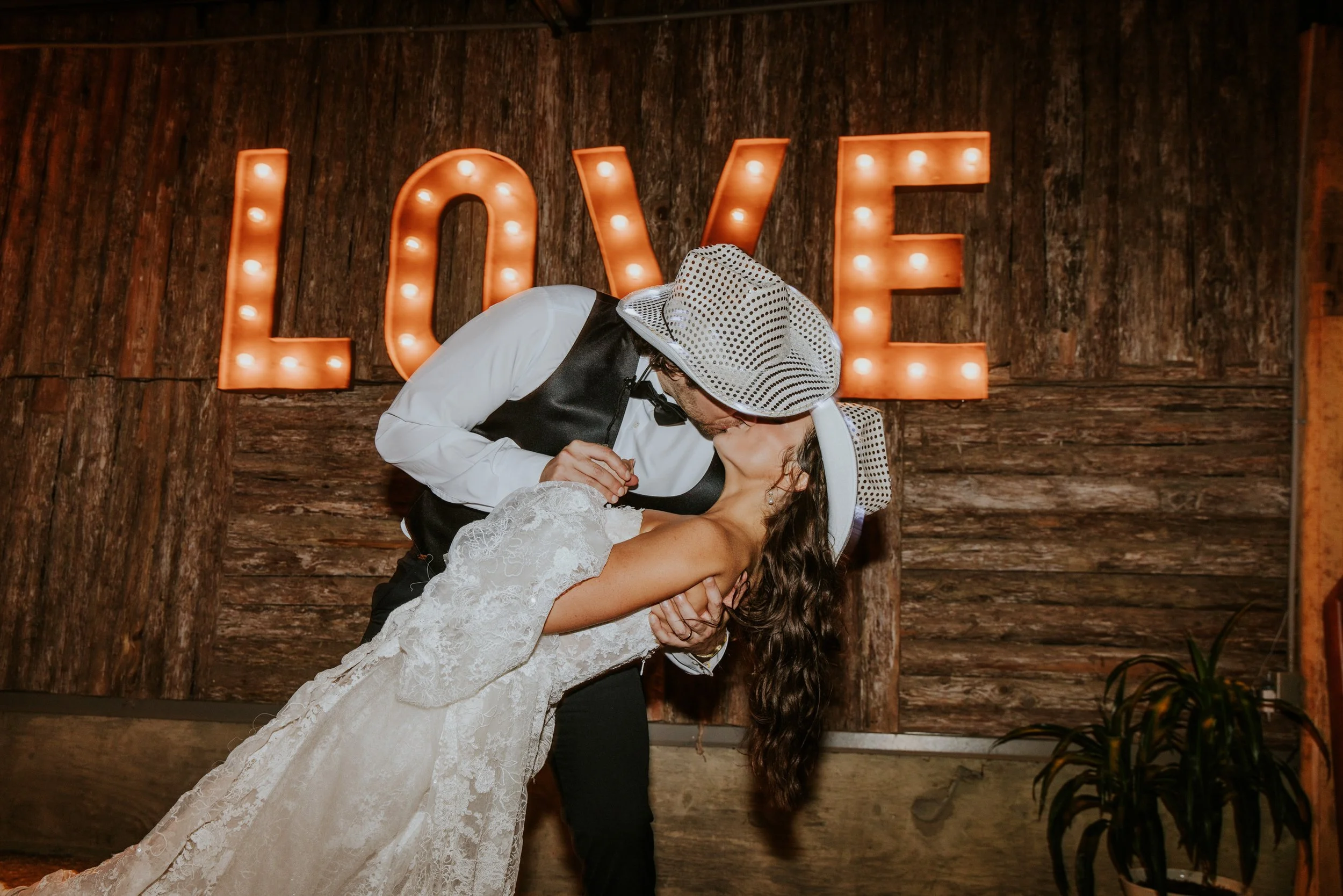 A couple sharing a kiss at a wedding reception; the man is wearing a cowboy hat and suspenders, the woman is in a white lace wedding gown, with a wooden wall and illuminated 'LOVE' sign in the background.