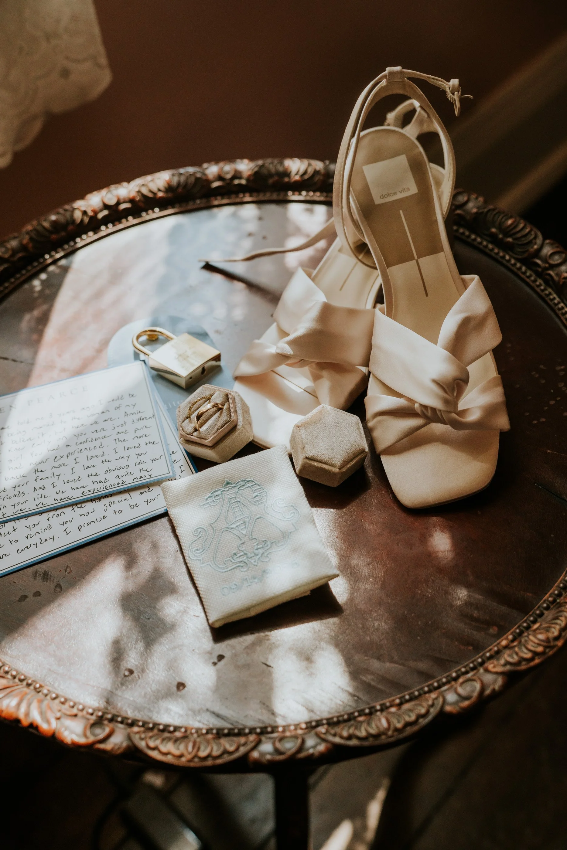 A pair of elegant cream-colored high-heeled shoes with bows on a vintage wooden table, along with a lock, a letter, a fabric pouch, and small hexagonal boxes, bathed in warm natural light.