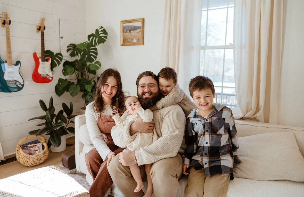 Family of five sitting on a cream-colored couch in a living room, smiling and playing together. The room has white walls, large windows with cream curtains, and musical guitars hanging on the wall.