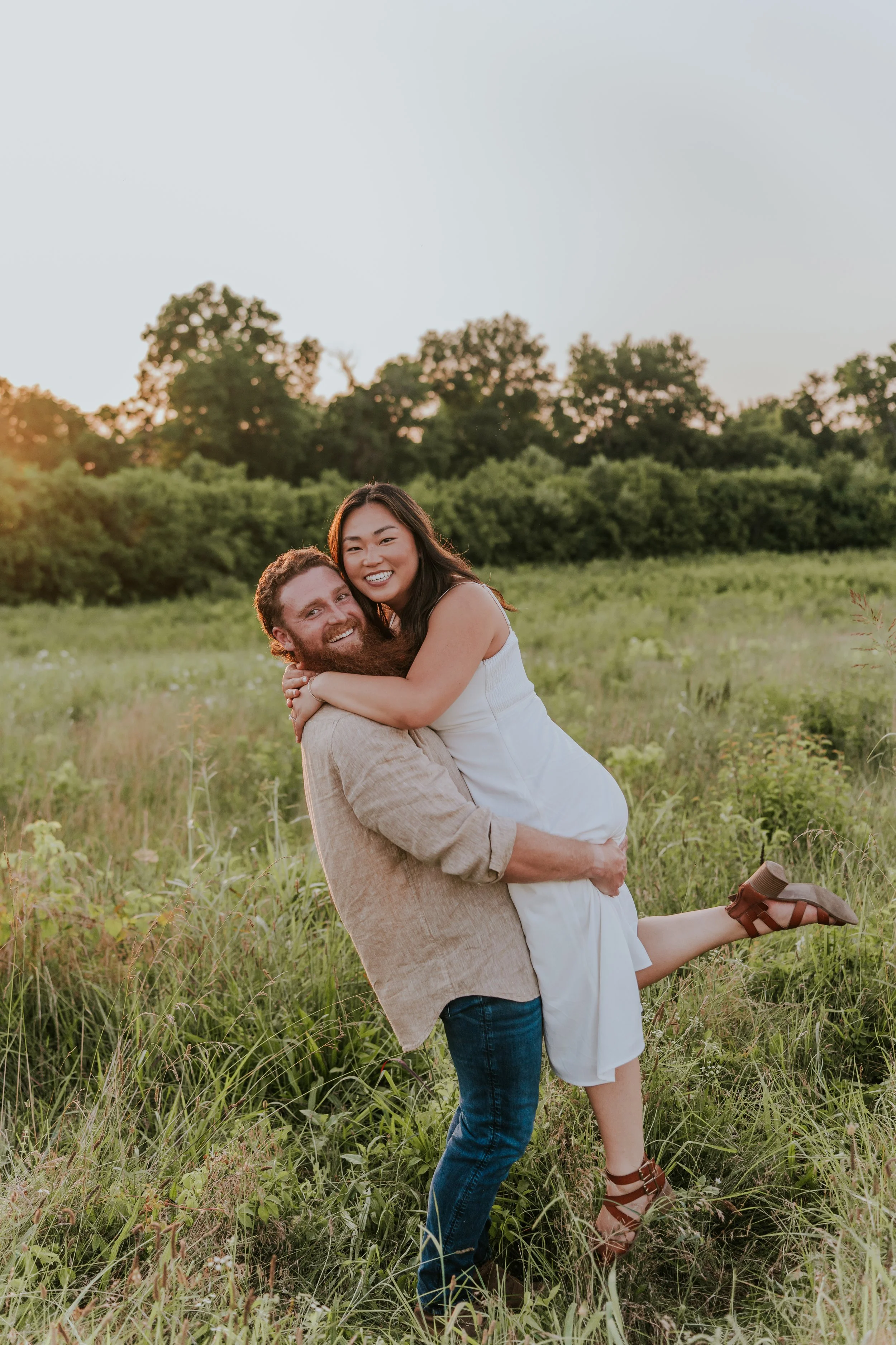 A couple outdoors in a grassy field, with the man lifting the woman, both smiling happily at sunset.