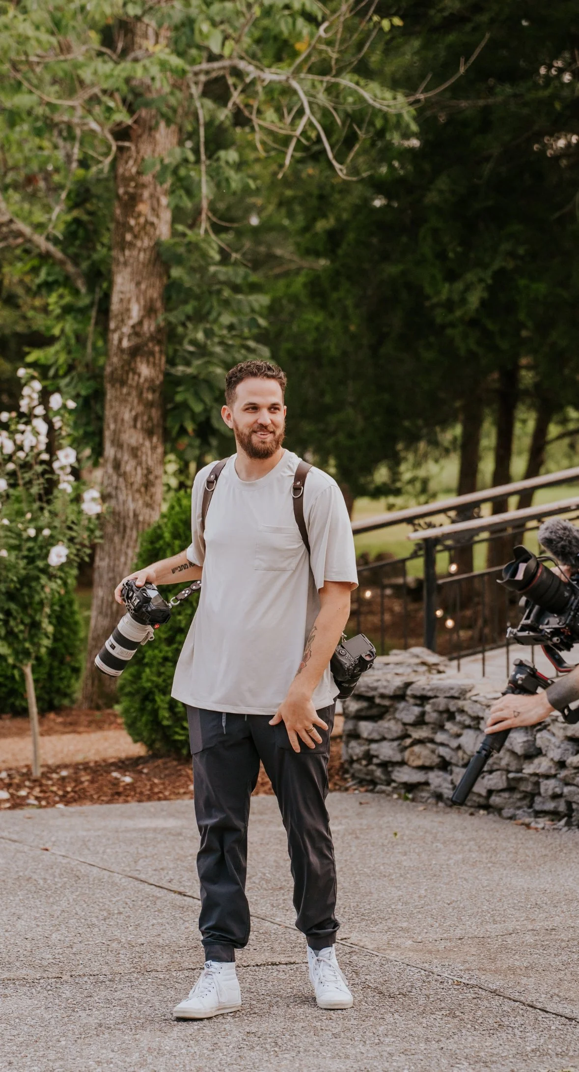 A man with a camera around his neck standing outdoors in front of trees and bushes, with a person operating a camera on a stabilizer to the right.