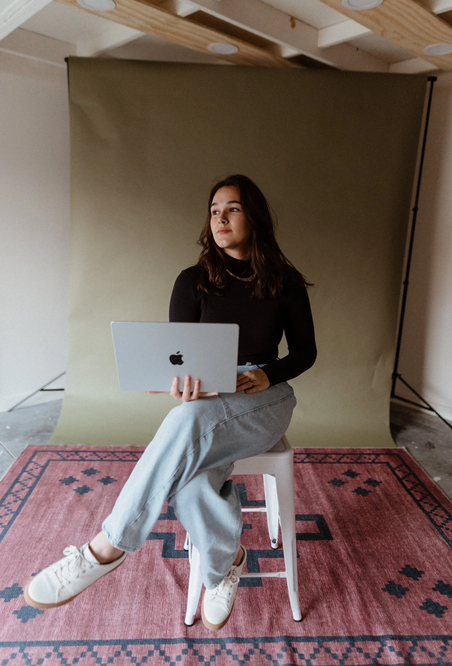 A young woman with long dark hair, wearing a black turtleneck and light gray jeans, sitting on a white stool on a pink patterned rug, holding a silver MacBook laptop, in a photography studio with a green backdrop and wood ceiling.