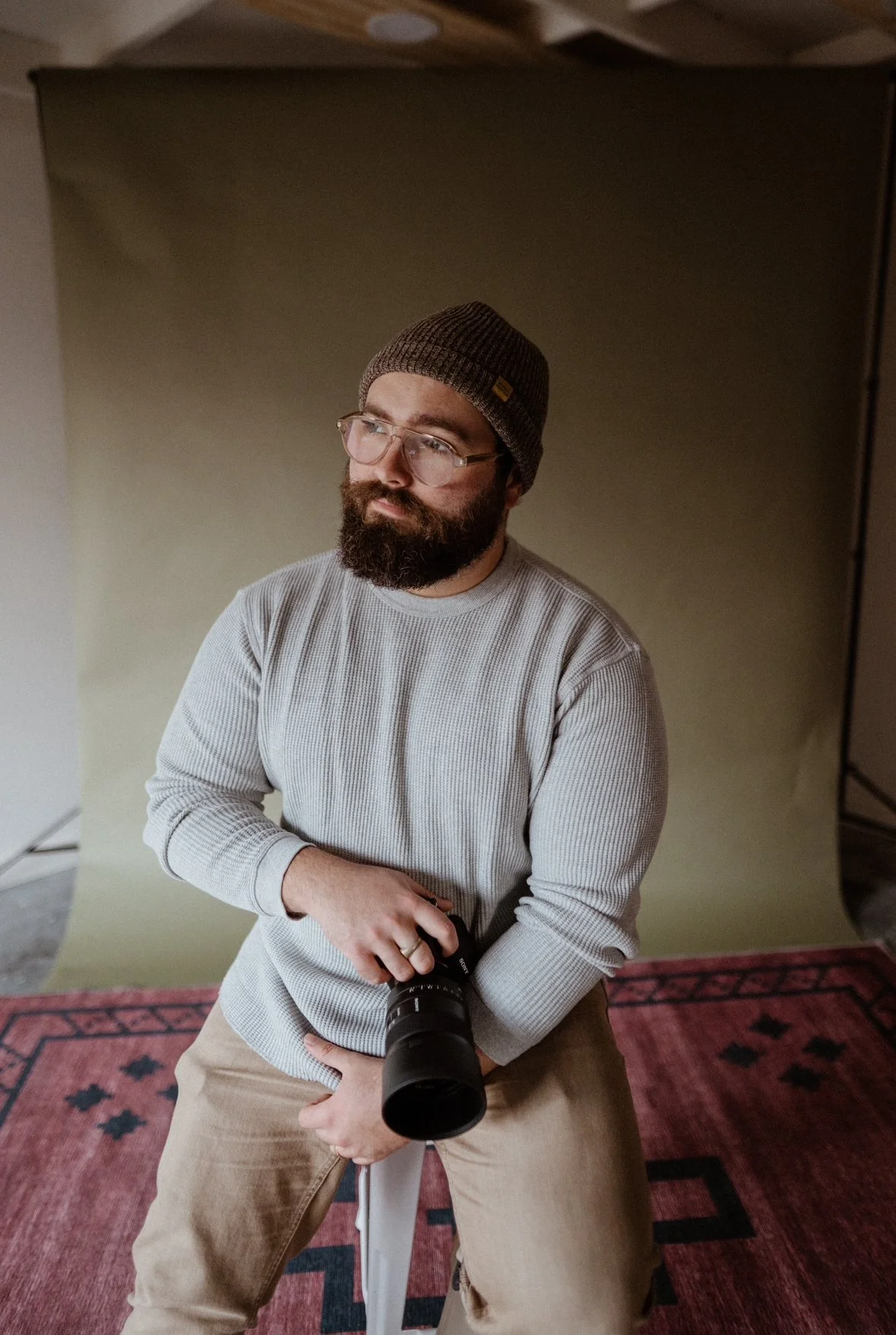 A man with a beard, glasses, and a beanie holding a camera, posing in front of a plain backdrop.