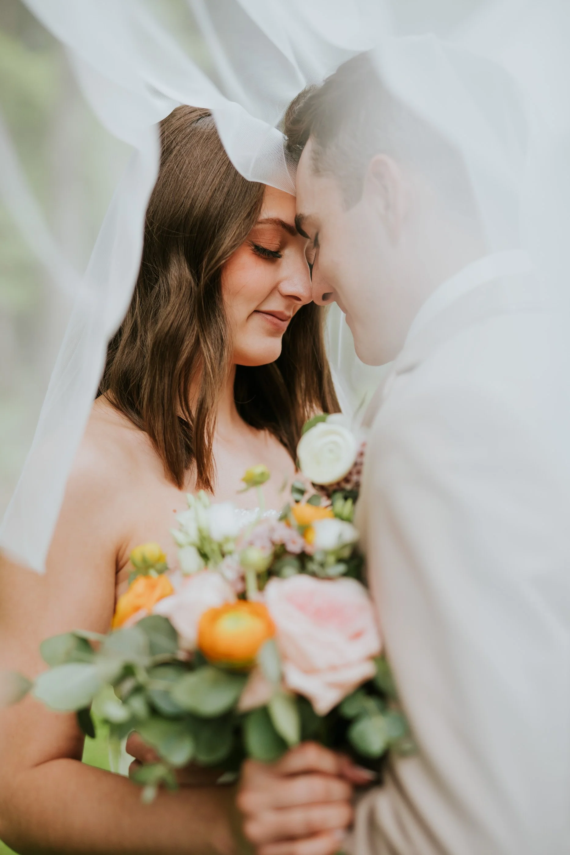 Close-up of a bride and groom with foreheads touching, holding a bouquet, under a sheer veil during their wedding.
