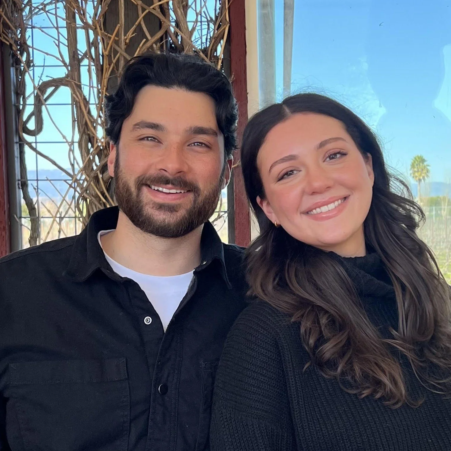 A smiling man with dark hair and a beard, wearing a black shirt, standing next to a smiling woman with long dark hair, wearing a black sweater, outdoors with a blue sky and palm trees in the background.