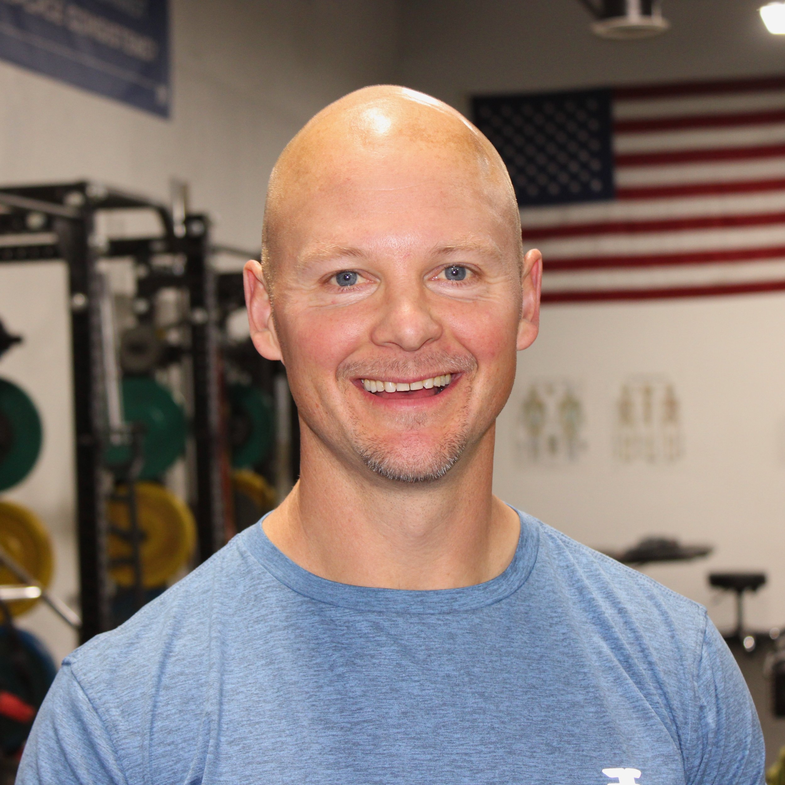A smiling man with a shaved head and light beard in a gym with weightlifting equipment and an American flag in the background.