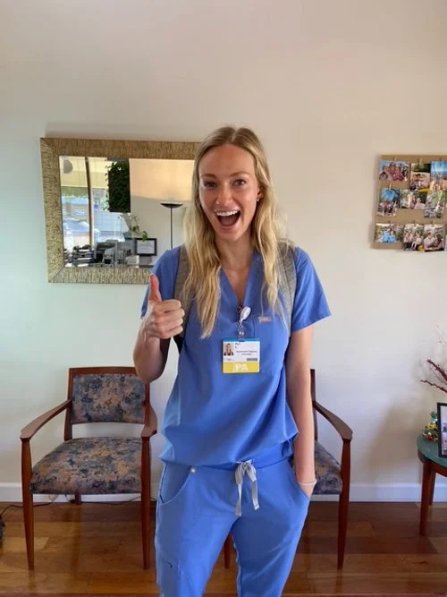 Young woman in blue scrubs smiling and giving a thumbs-up inside a room with a mirror, framed photos, and chairs.