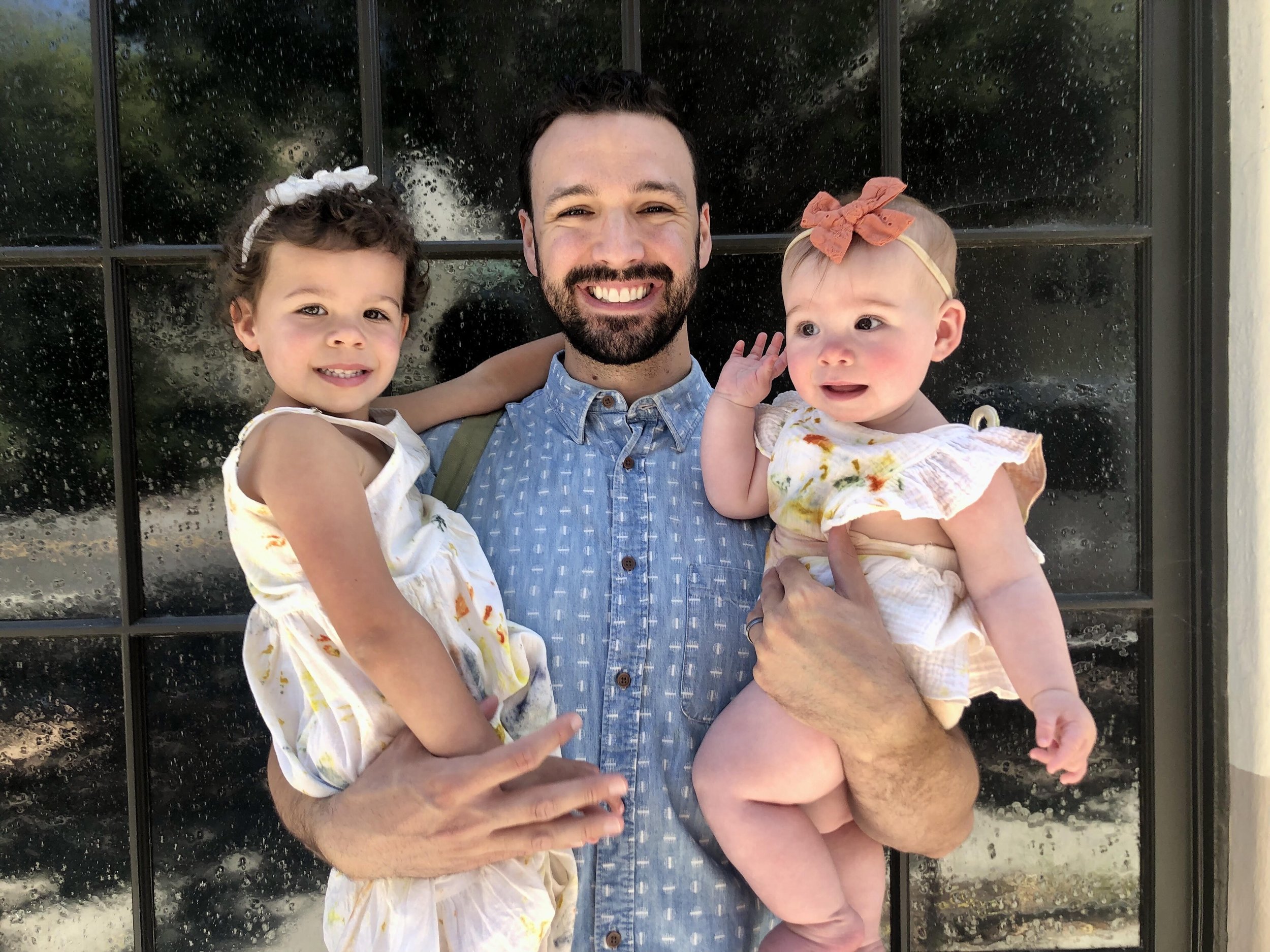 A smiling man holding two young girls, one on each arm, standing in front of a black window with water drops. The man has a beard and is wearing a blue button-up shirt. The girl on the left has curly hair with a white headband and is wearing a white dress with orange and yellow patterns. The girl on the right has light hair with a pink bow and is wearing a white dress with colorful stains.
