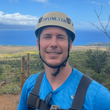 Man wearing safety helmet and blue sports shirt, smiling outdoors with a scenic landscape and ocean in the background.