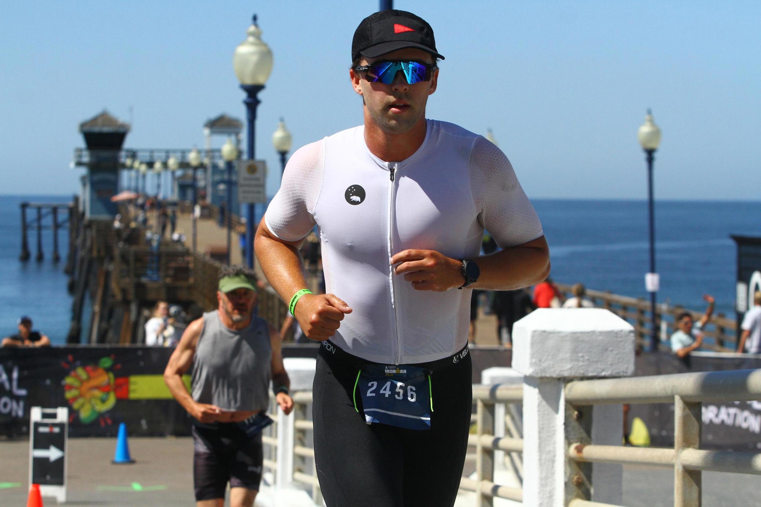 Man running in a race near a pier by the ocean, wearing sunglasses, a black cap, a white athletic shirt, and black shorts, with race bib number 2456.