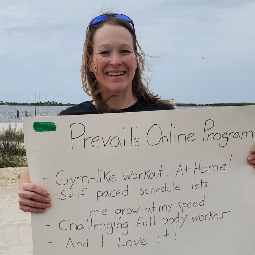 A woman smiling on a beach holding a whiteboard with handwritten text about Prevail's online program, which offers gym-like workouts at home with a self-paced schedule for full body workouts.