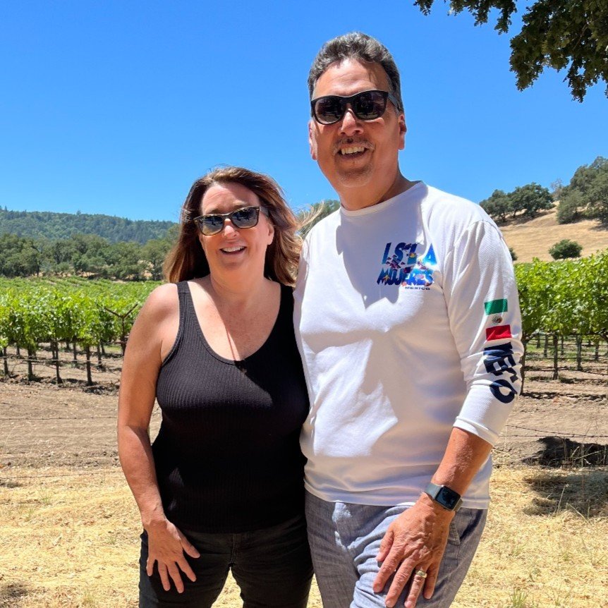 A smiling man and woman standing together outdoors in front of a vineyard, under a clear blue sky, with greenery and hills in the background.