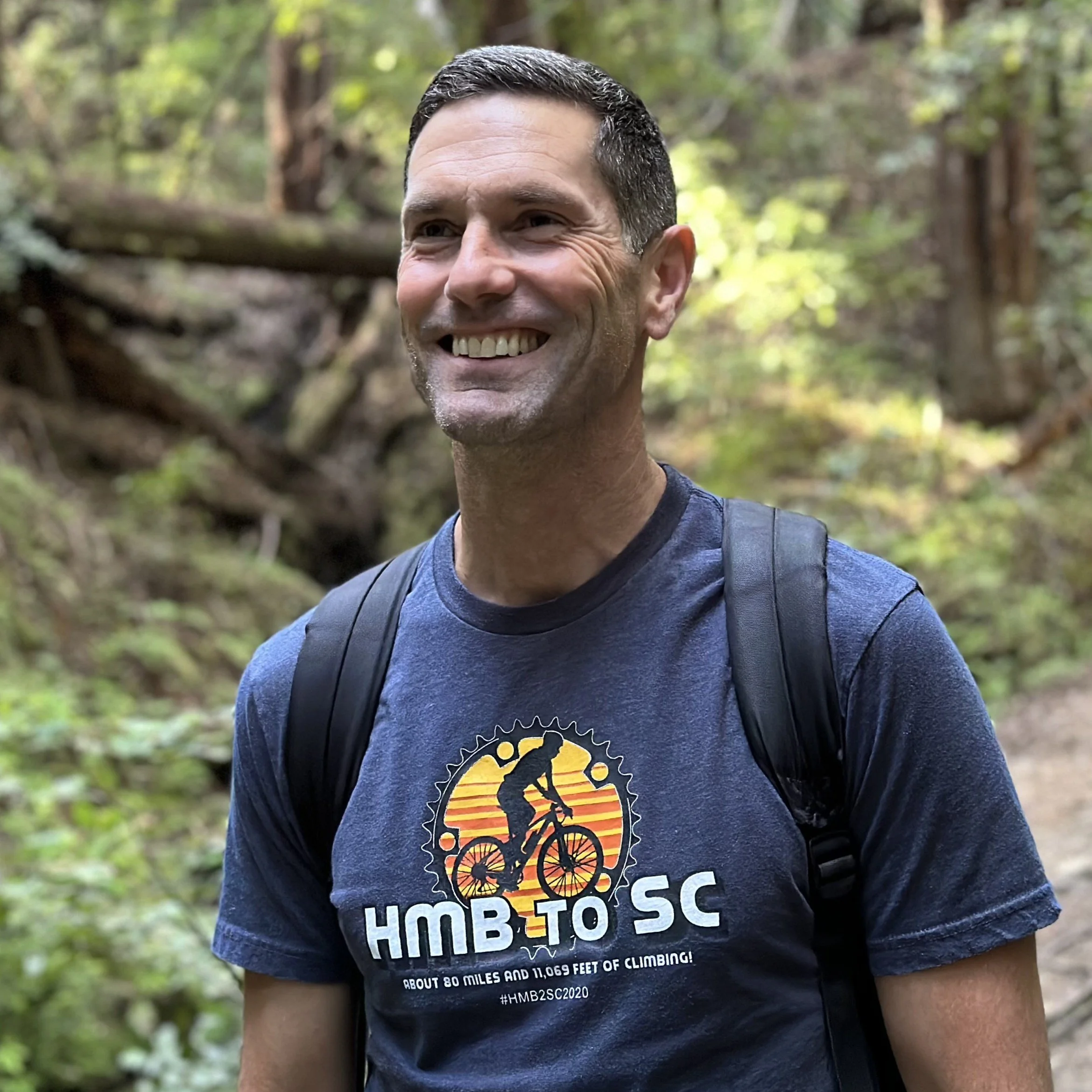 A smiling man wearing a blue t-shirt with a mountain biking graphic and the text 'HMB to SC,' standing outdoors in a forested area.
