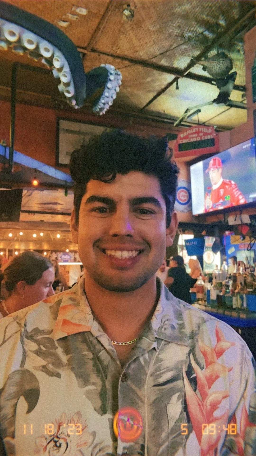 A young man with dark hair and a floral shirt smiling in a bar or restaurant with a busy background including a TV, chandeliers, and people.