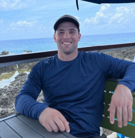 Man smiling sitting at an outdoor table near the beach, with ocean and rocky shoreline in the background.