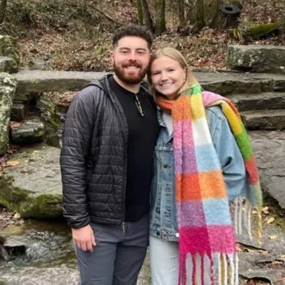 A smiling couple standing outdoors near a small stream, surrounded by trees and rocks. The man has a beard, wearing a black jacket, and the woman has blonde hair, wearing a colorful striped scarf and a denim jacket.