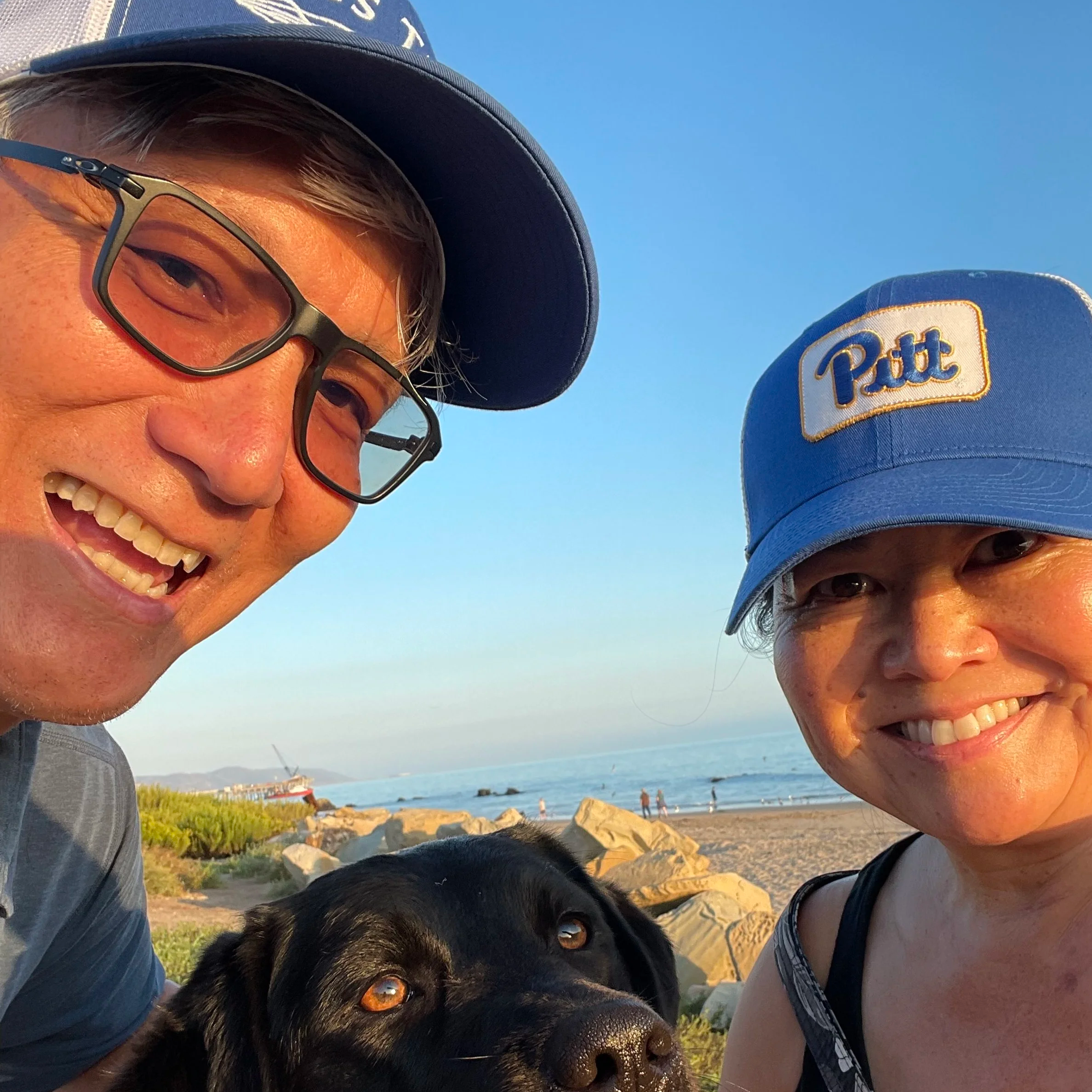 Two smiling people wearing blue hats and a black dog with a beach and ocean in the background during sunset.