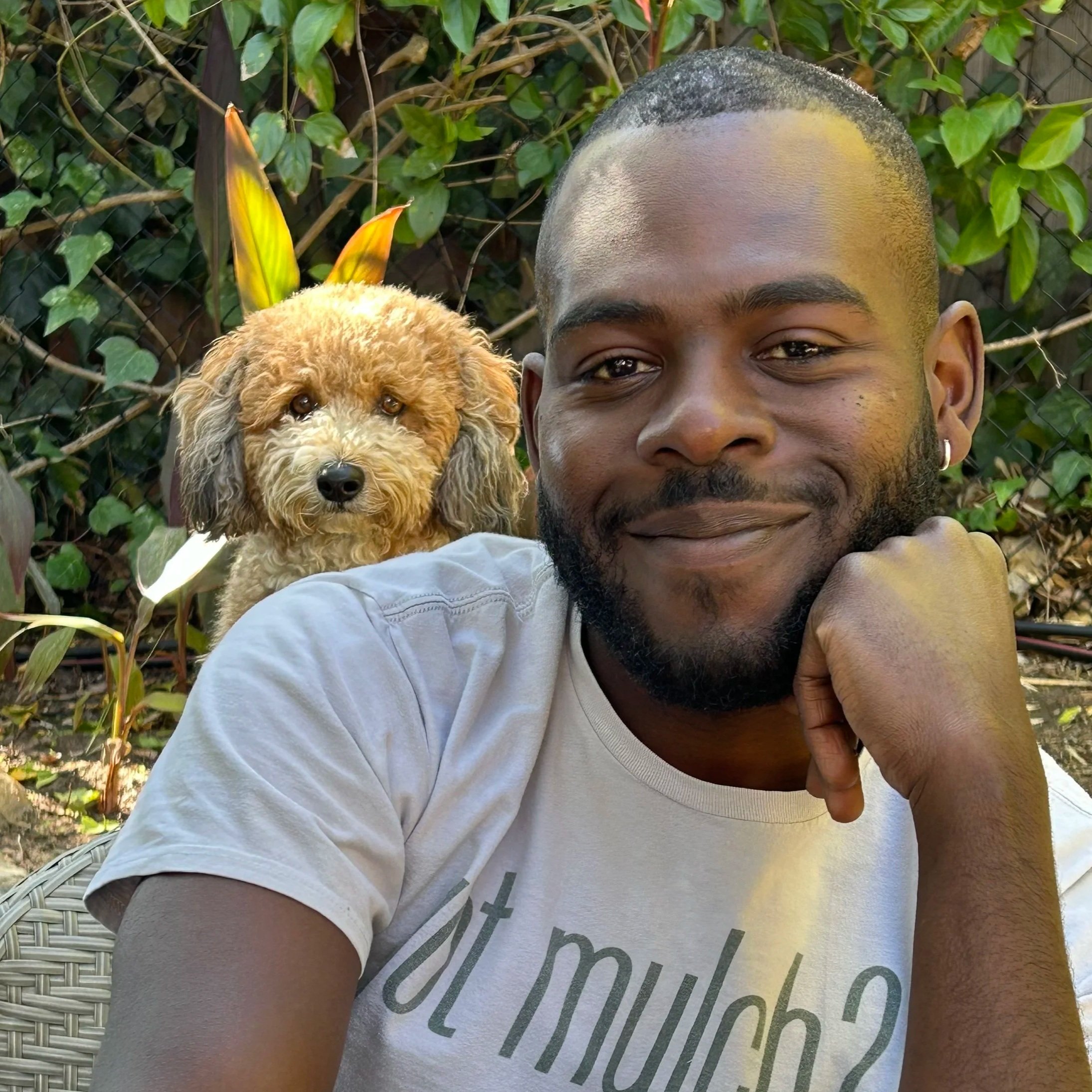 A man with a beard and earrings smiling at the camera with a small fluffy brown dog sitting behind him, surrounded by green plants.