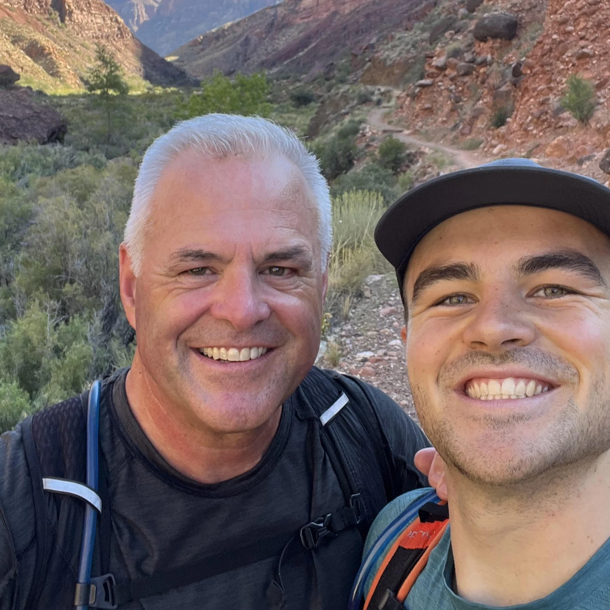 Two smiling men taking a selfie outdoors in a mountainous area with green vegetation and rocky terrain.