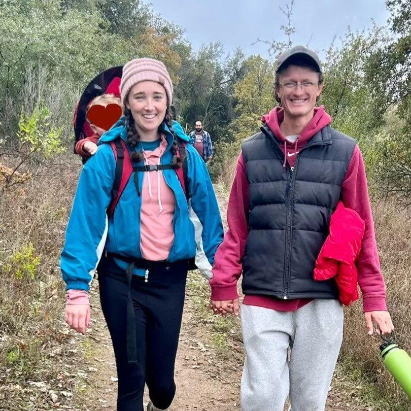 A young couple is hiking on a trail in a wooded area, holding hands and smiling at the camera. The woman is wearing a pink beanie, blue jacket, and black pants, with a backpack. The man is wearing a gray cap, pink hoodie, black puffer vest, and gray sweatpants, holding a green water bottle.