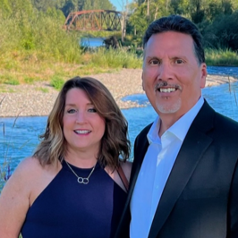 A smiling couple outdoors by a river, with a bridge and trees in the background.