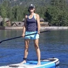 A woman standing on a paddleboard on a lake, holding a paddle, with trees and a dock in the background.