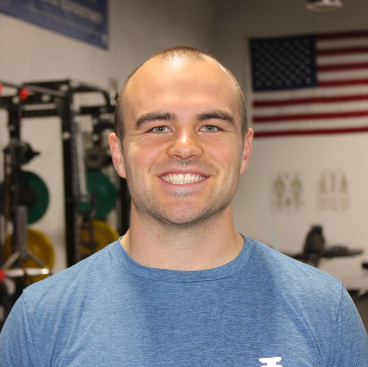 A smiling man with a shaved head wearing a blue athletic shirt in a gym with weightlifting equipment and an American flag in the background.