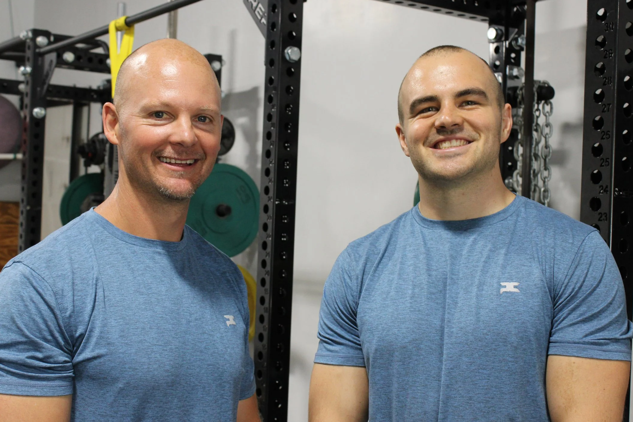 Two smiling men in blue t-shirts standing in a gym near weightlifting equipment.
