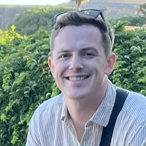 A young man with short, light brown hair smiling outdoors, wearing a striped shirt and sunglasses on his head, with greenery and trees in the background.