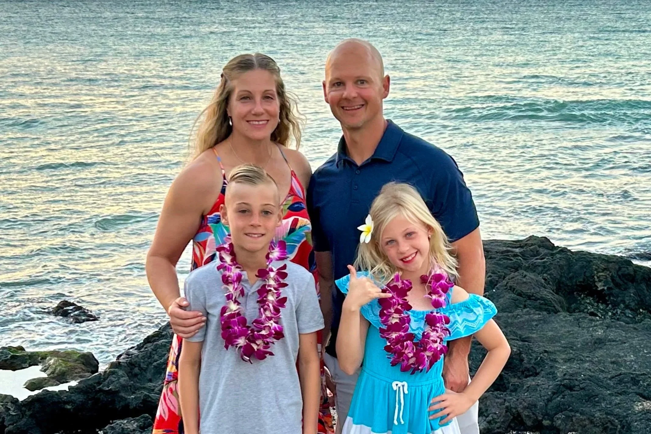 Family of four on rocks at the beach with ocean in the background, all wearing leis and smiling, during sunset.
