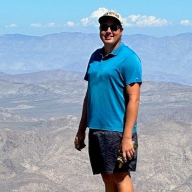 Man wearing a blue polo shirt, sunglasses, and a cap standing outdoors with a mountain landscape in the background.
