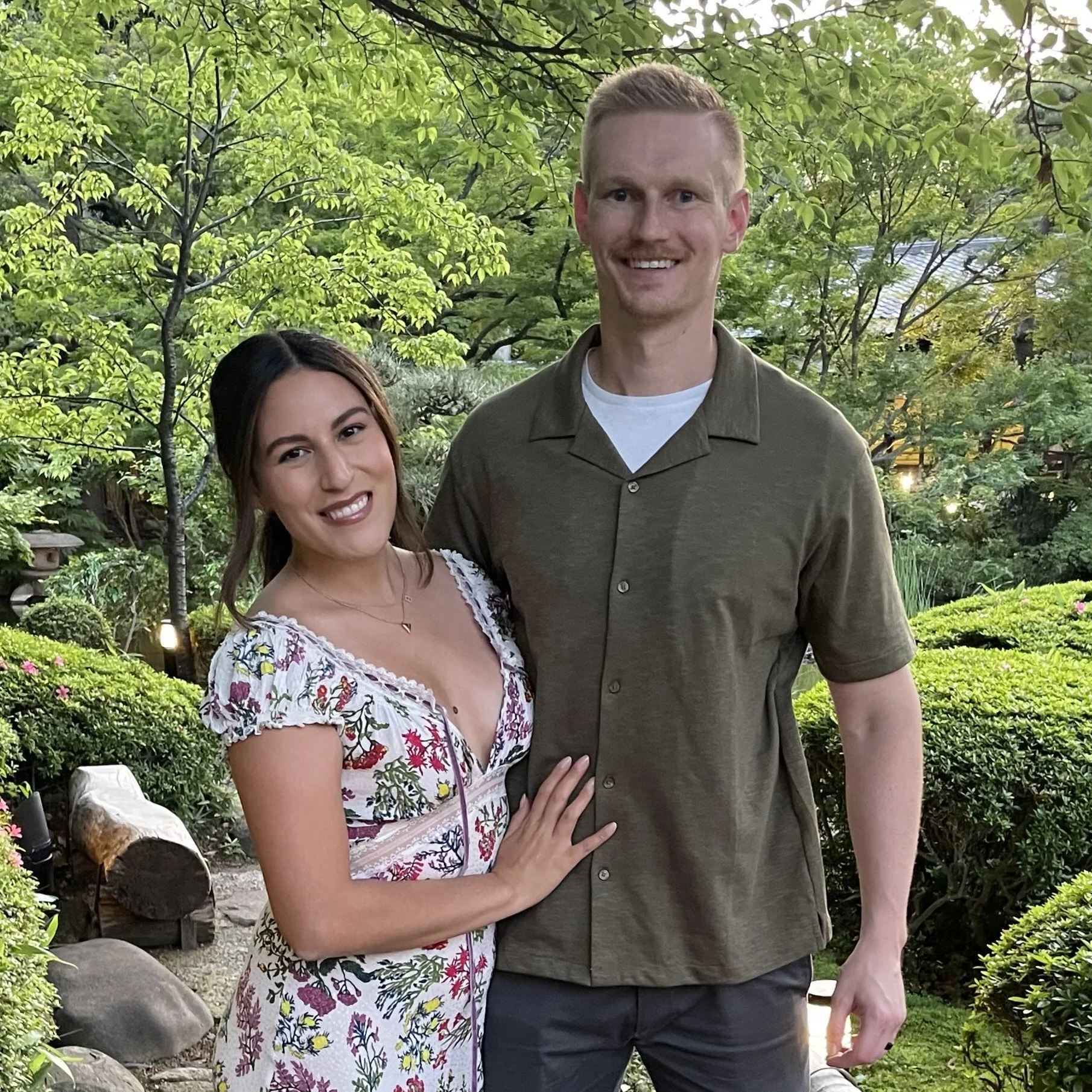 A smiling woman and a smiling man standing close together outdoors in a lush garden with green trees and bushes.