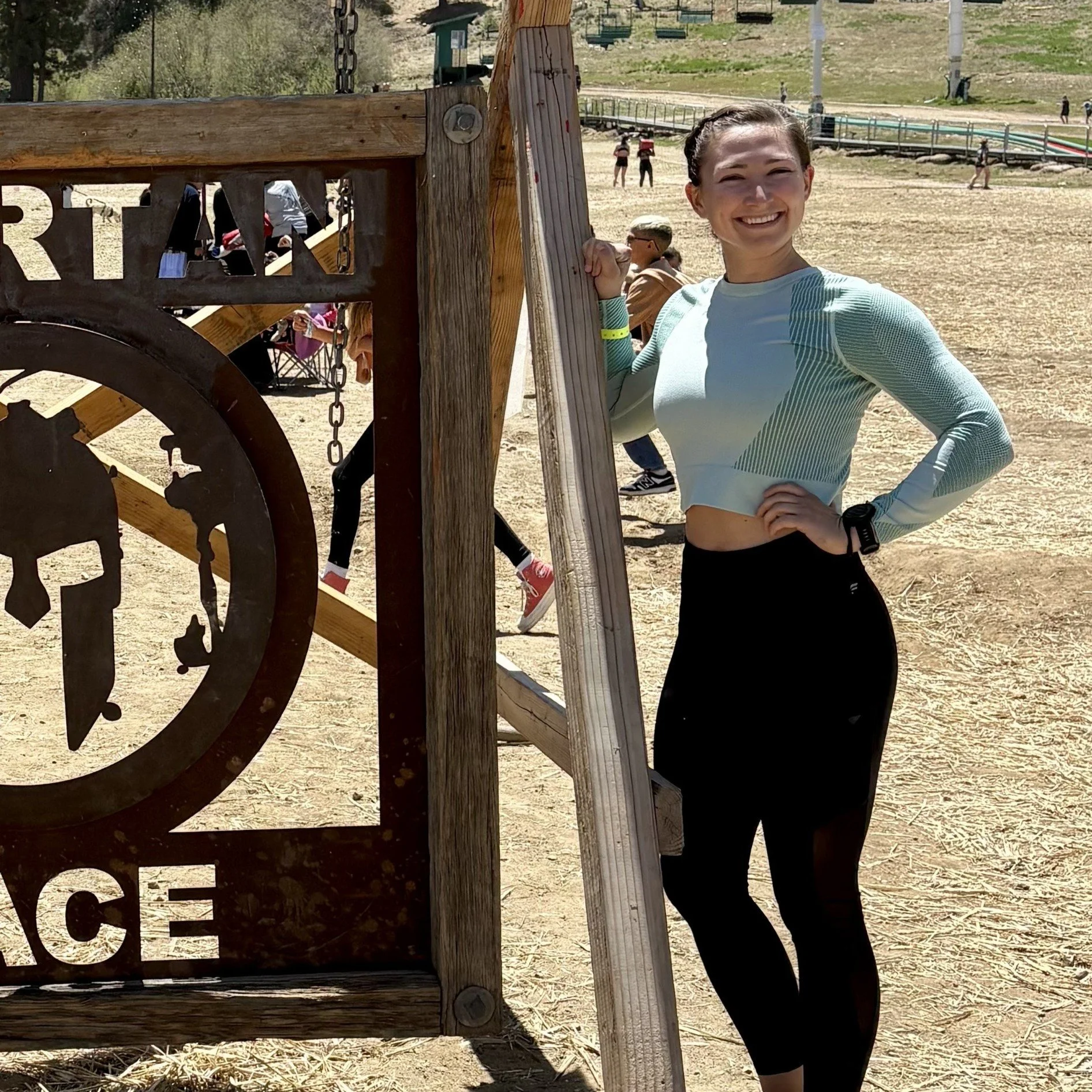 A young woman smiling and posing outdoors at a park or playground, standing next to a wooden structure with a metal sign that has a clock and some cut-out designs.