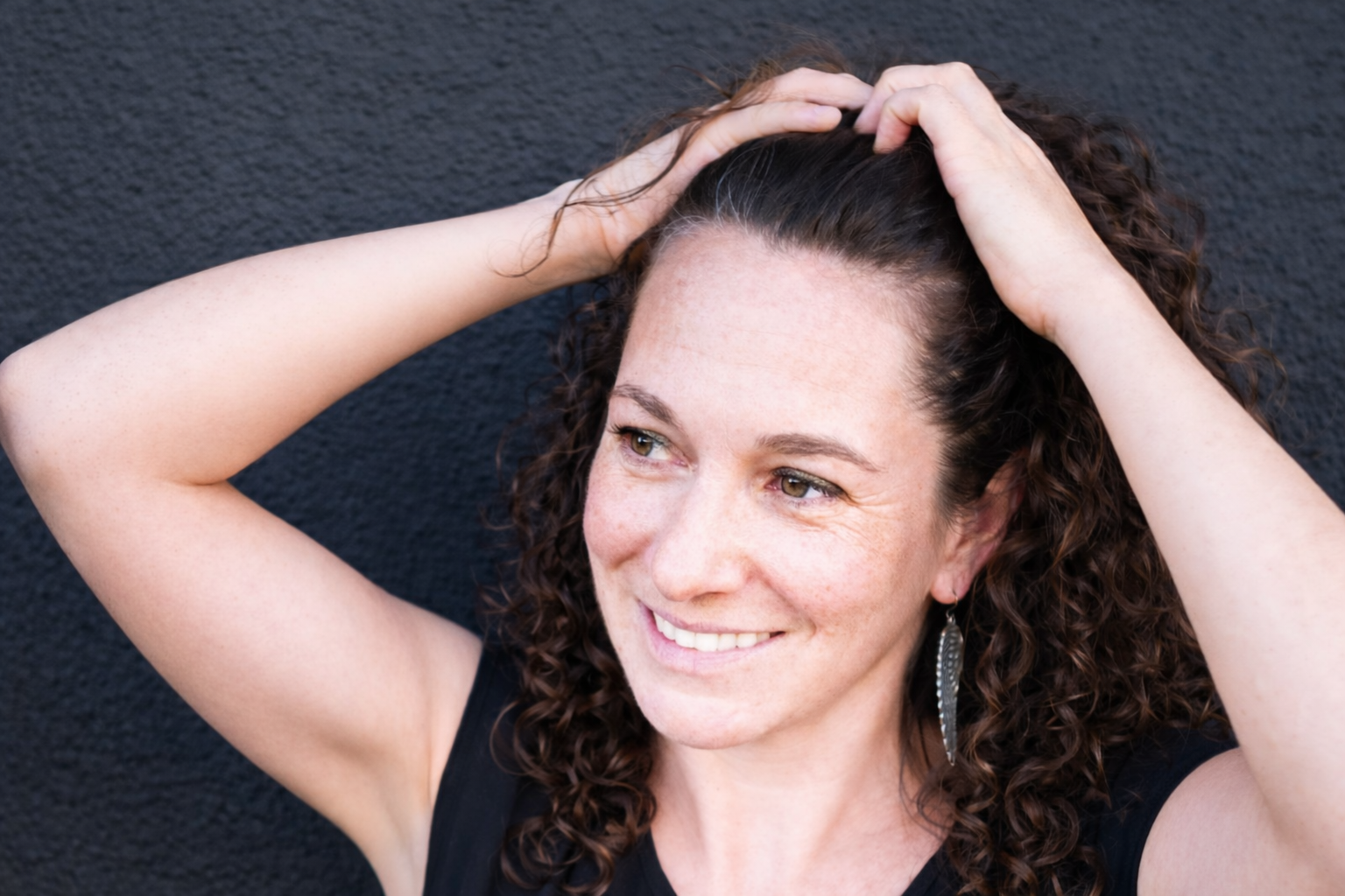 A woman with curly brown hair smiling and looking to her right, holding her hair up with both hands, standing against a black textured wall.