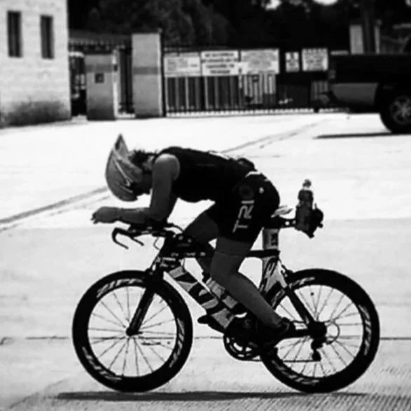 A person wearing a helmet and cycling gear is riding a bicycle on a paved road with a gated area and building in the background.