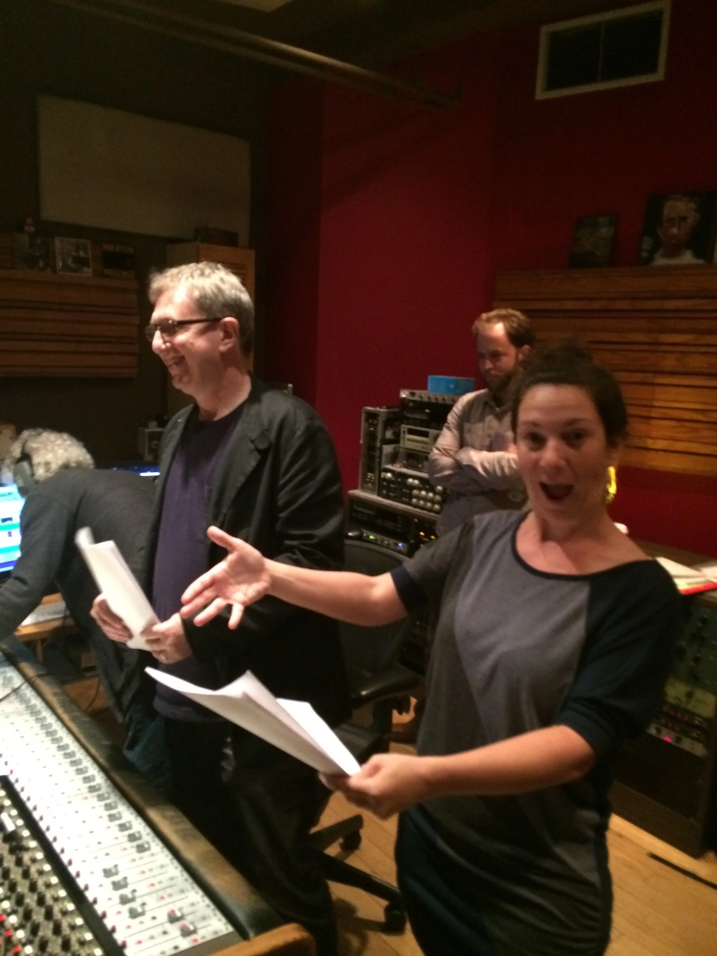 Three people in a recording studio, two women and one man, appear to be working or discussing music or sound. The woman in the foreground is holding a paper and gesturing, the man is smiling with his arms crossed, and the other woman is looking at a 
